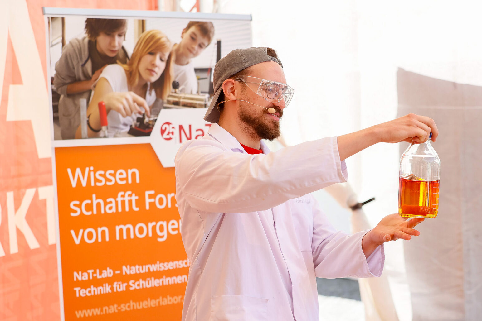 Scienziati con liquido arancione davanti a un roll-up "La conoscenza crea la ricerca di domani".