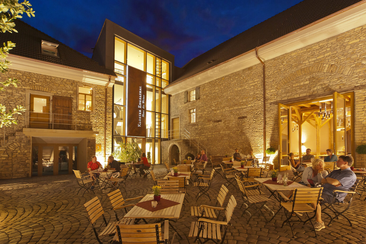 Historische Bausubstanz und moderne Architektur, im Vordergrund ein Außenbereich mit Menschen, die an Holztischen sitzen.