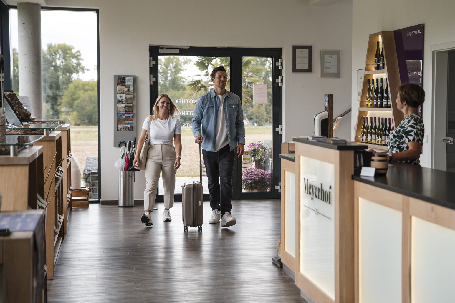 Eine junge Frau und ein junger Mann mit Trolley kommen in einen Raum, rechts ist ein Counter mit dem Schriftzug Meyerhof und eine Frau, die dahinter steht.