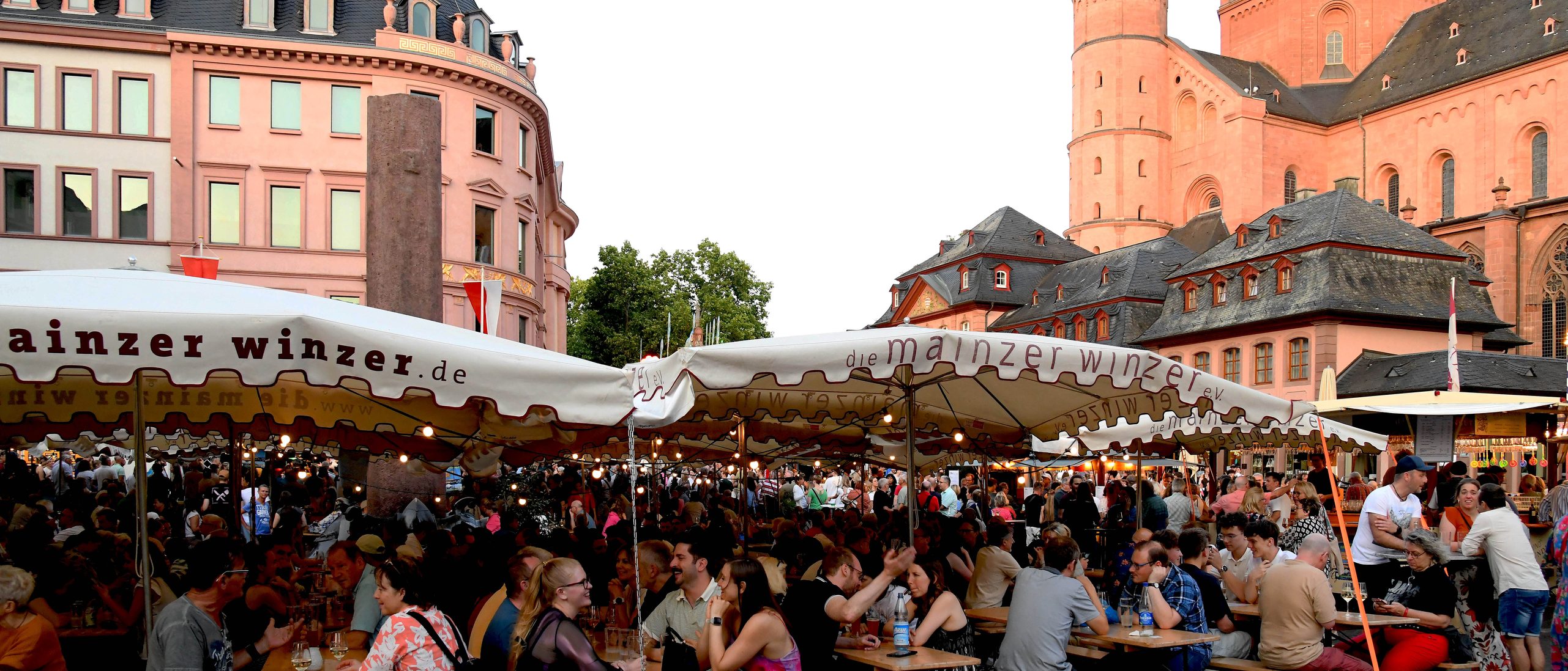 Au marché, des gens sont assis sur des bancs de brasserie, à l'ombre des parasols des vignerons de Mayence, à l'occasion de la fête de la Saint-Jean de Mayence. À droite, en arrière-plan, on aperçoit la cathédrale.
