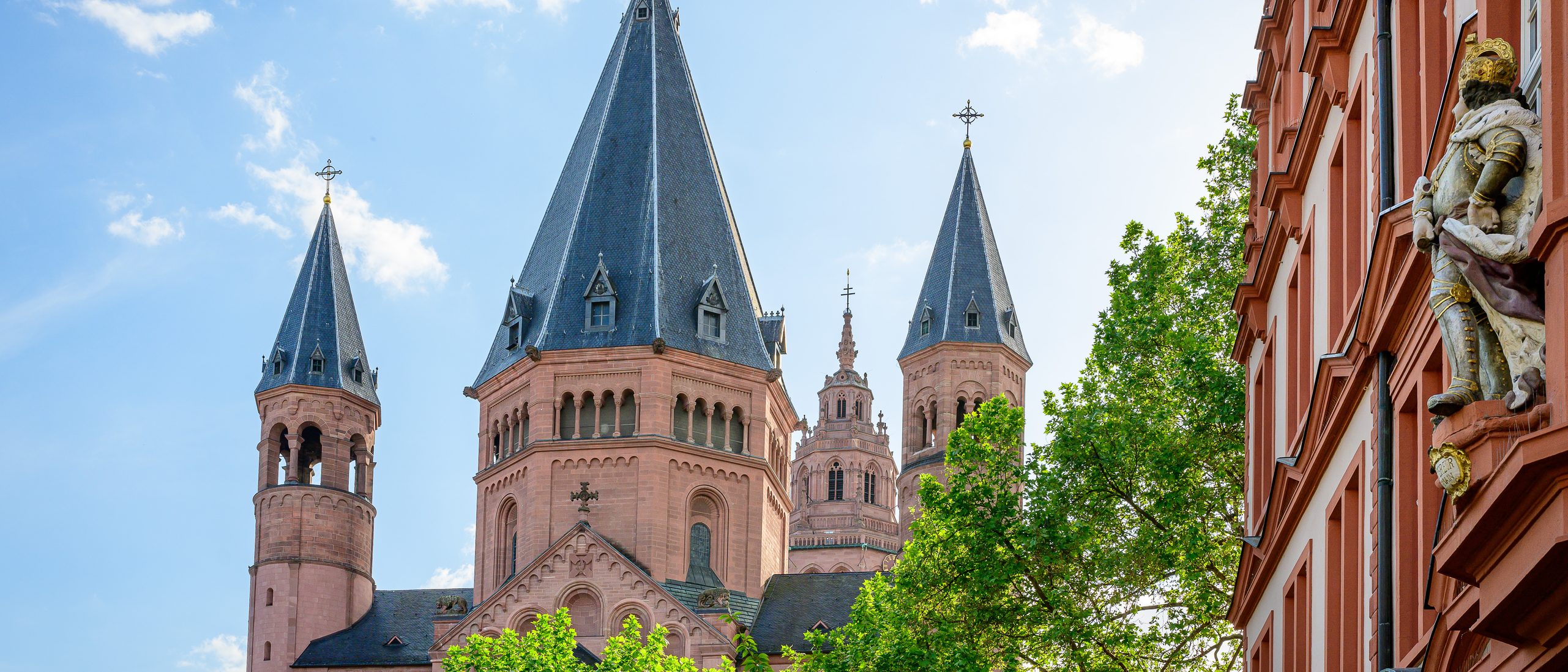 In the center is the east choir of the cathedral in red sandstone, in front of it green treetops, on the right the Haus zum Römischen Kaiser, white with red elements
