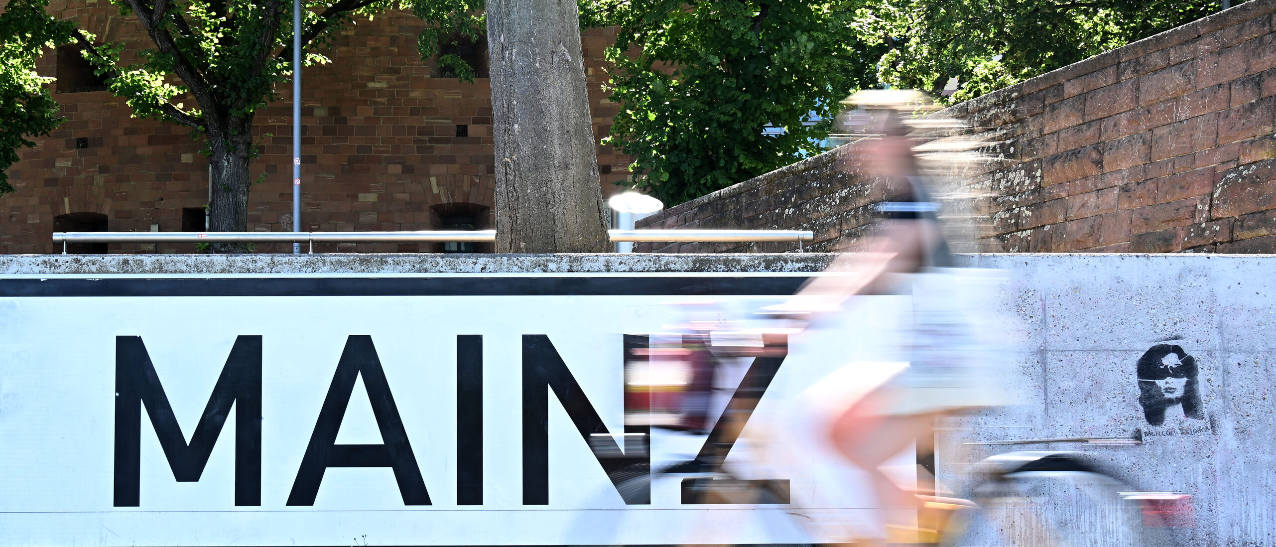 Lettering MAINZ in capital letters, black on white on a gray wall, in front of it a cyclist (blurred)
