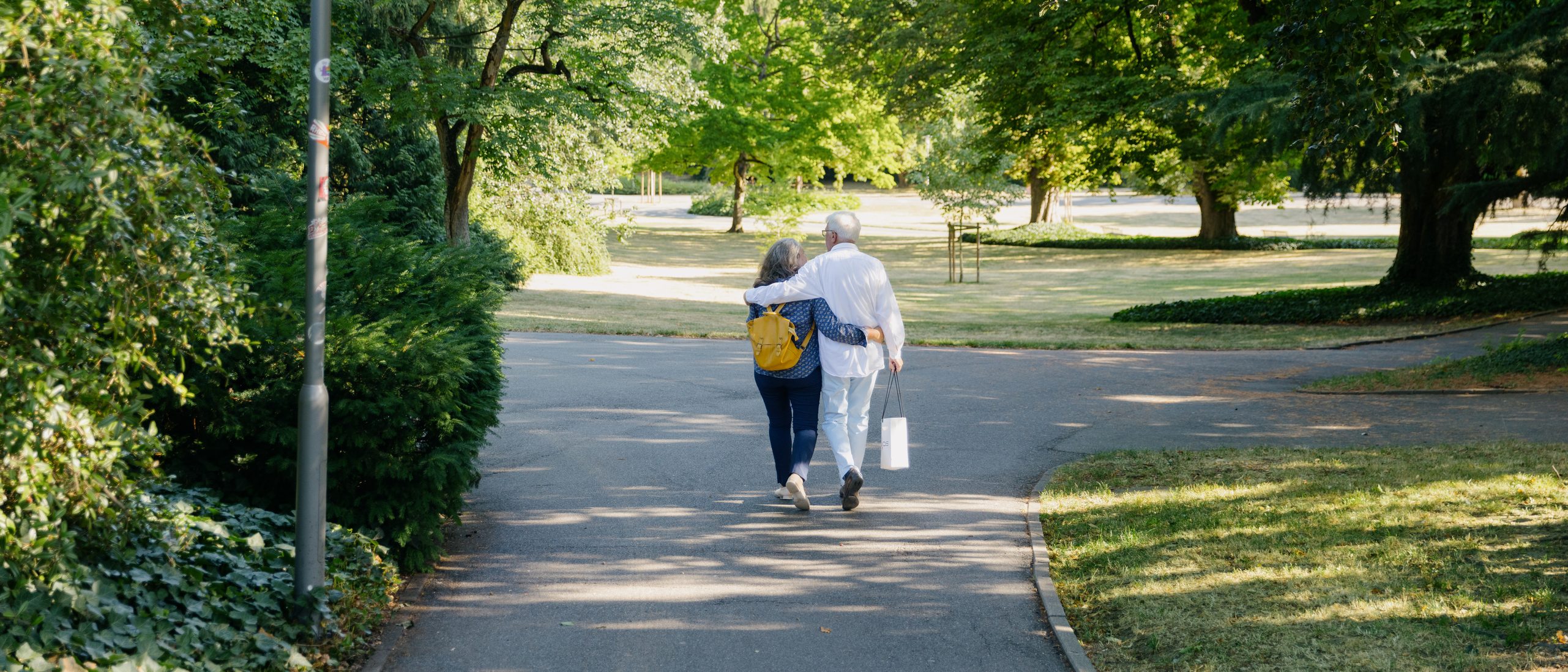 Una mujer y un hombre pasean por el parque de la ciudad; se les ve de espaldas, rodeados de árboles, un prado y una farola.