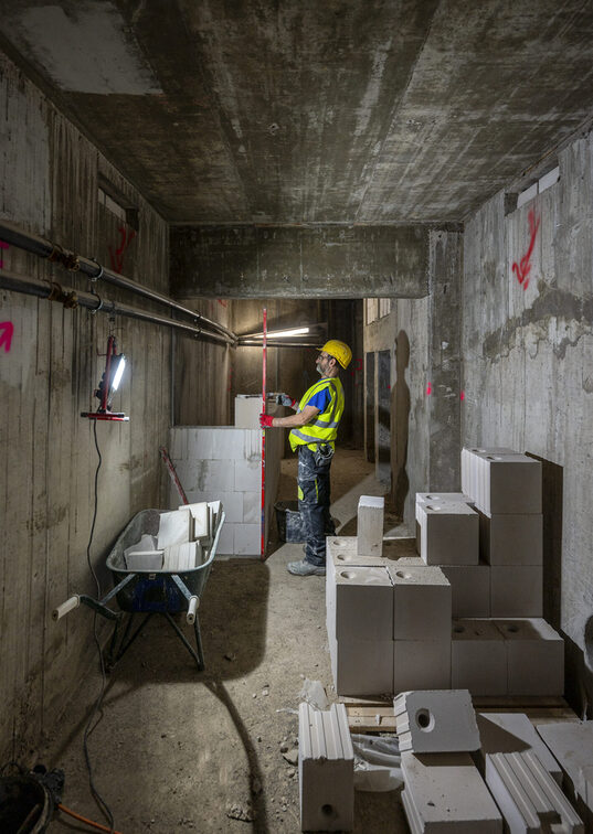 Lavoratori edili in uno stretto corridoio, circondati da elementi di cemento.