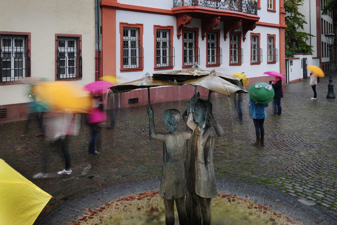 Menschen mit Schirmen um den Mädchenbrunnen am Ballplatz