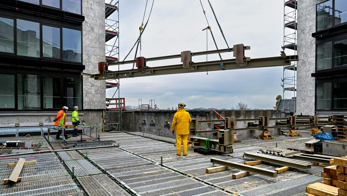 Drei Bauarbeiter auf der Baustelle des Rathauses nehmen ein Stahlteil entegegen.