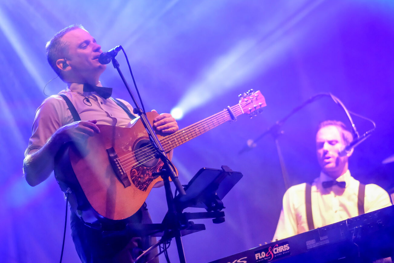 Zwei Männer auf einer Bühne, einer hat eine Gitarre umgehängt und steht am Mikrofon, der andere sitzt am Keyboard.