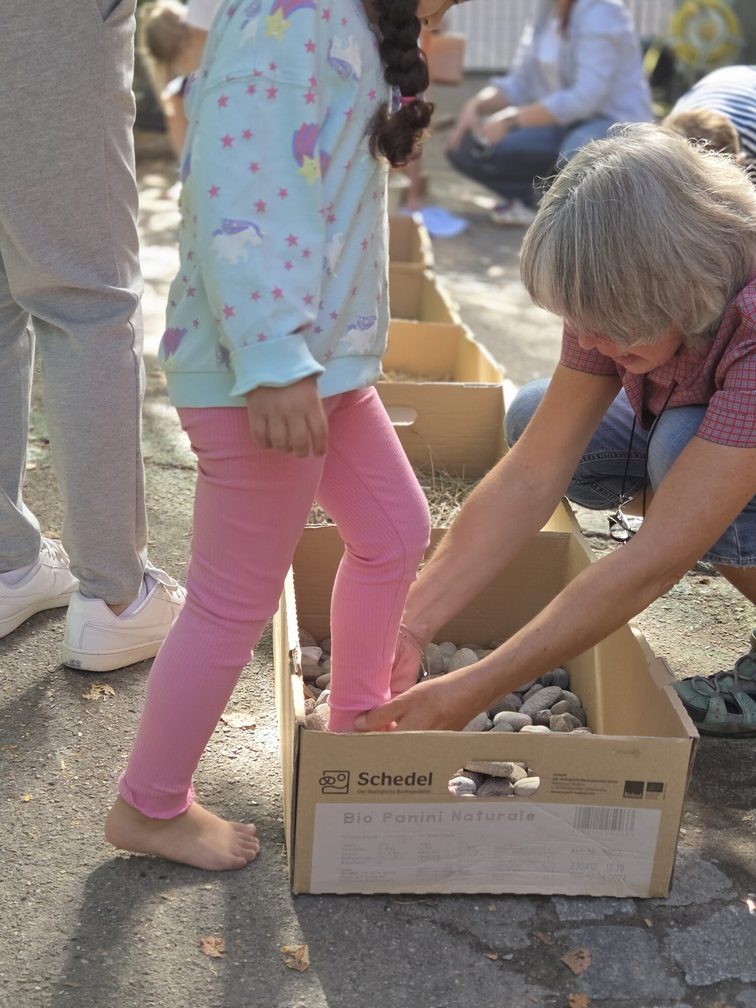 Gambe di bambini che, aiutati da un’educatrice, salgono su una cassetta di un percorso a piedi nudi
