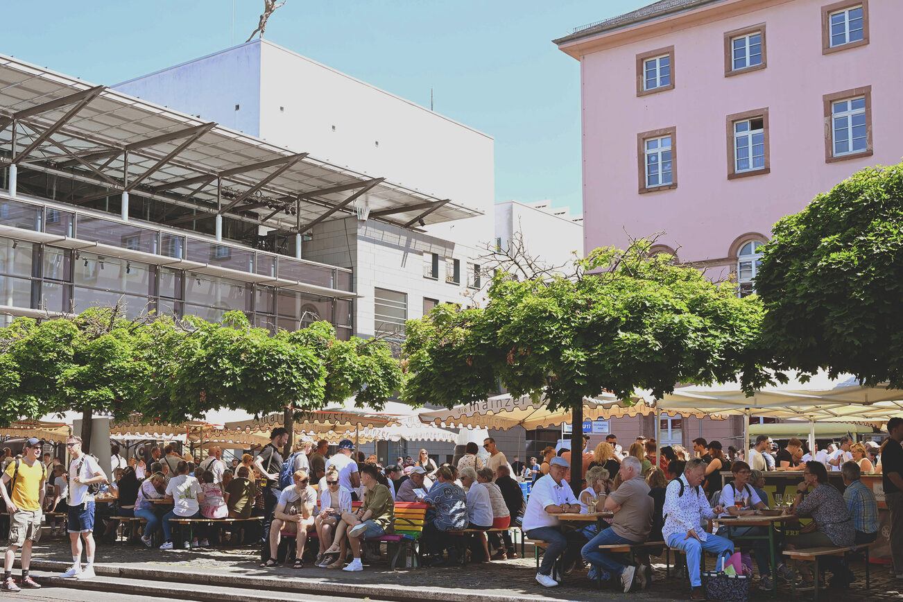 Besucher:innen des Marktfrühstücks sitzen auf Bänken am Tritonplatz