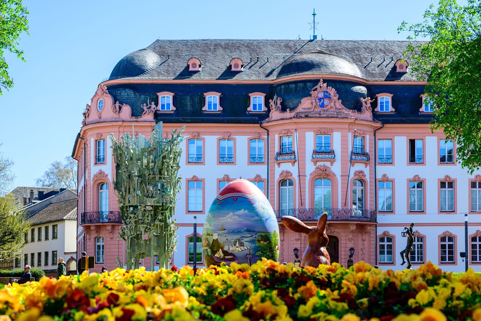 In the foreground a flower bed with predominantly yellow blossoms, to the left the carnival fountain, in the middle a huge, painted Easter egg and next to it a hare figure from behind in front of a large aristocratic palace (Osteiner Hof).