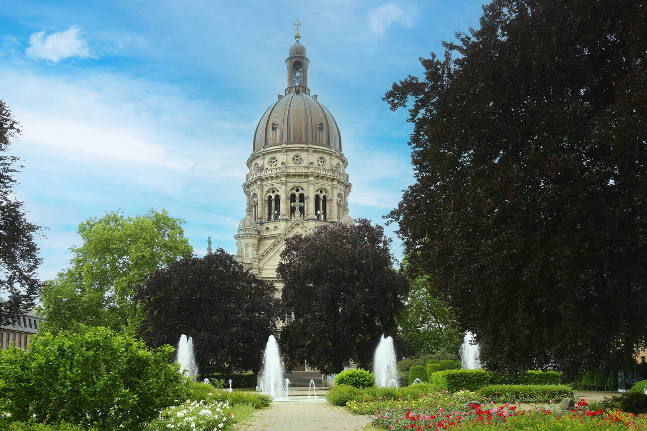 Christuskirche im Frühling, umrahmt von Bäumen, im Vordergrund der Brunnen mit den sprudelnden Fontänen