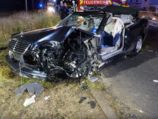 Demoliertes Unfallfahrzeug, Einsatzkräfte im Hintergrund.