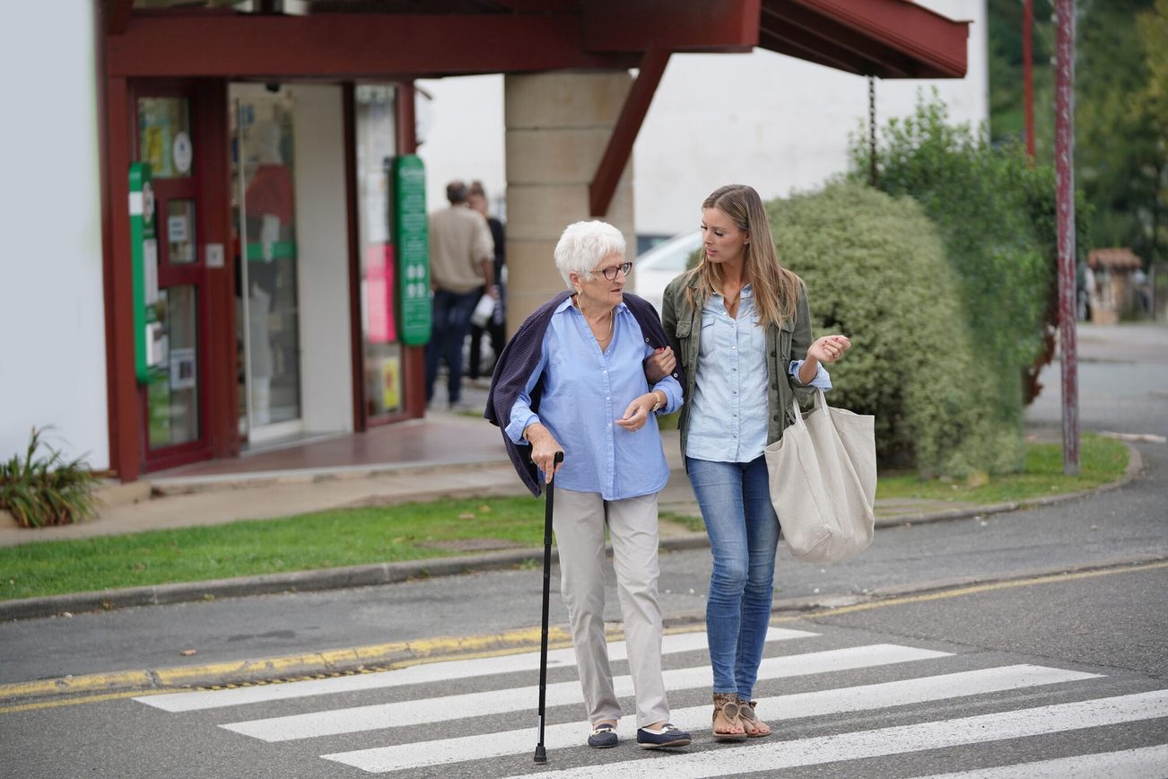 Eine Frau begleitet eine Seniorin mit Gehhilfe über einen Zebrastreifen