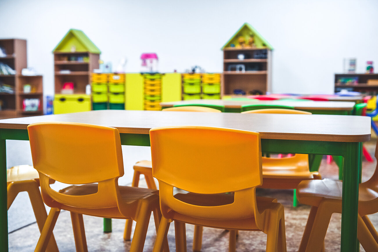 كراسي بلاستيكية صفراء على طاولة في المقدمة في مركز للرعاية النهارية، وفي الخلفية أرفف ذات حجرات صفراء وخضراء.