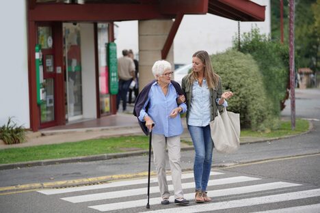 Eine Frau begleitet eine Seniorin mit Gehhilfe über einen Zebrastreifen